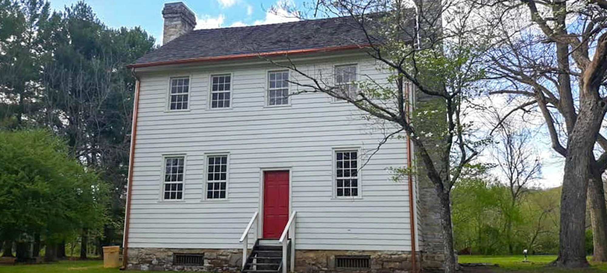 Historic two-story white wooden house with a red door, stone foundation, surrounded by lush green trees and grass under a partly cloudy sky.