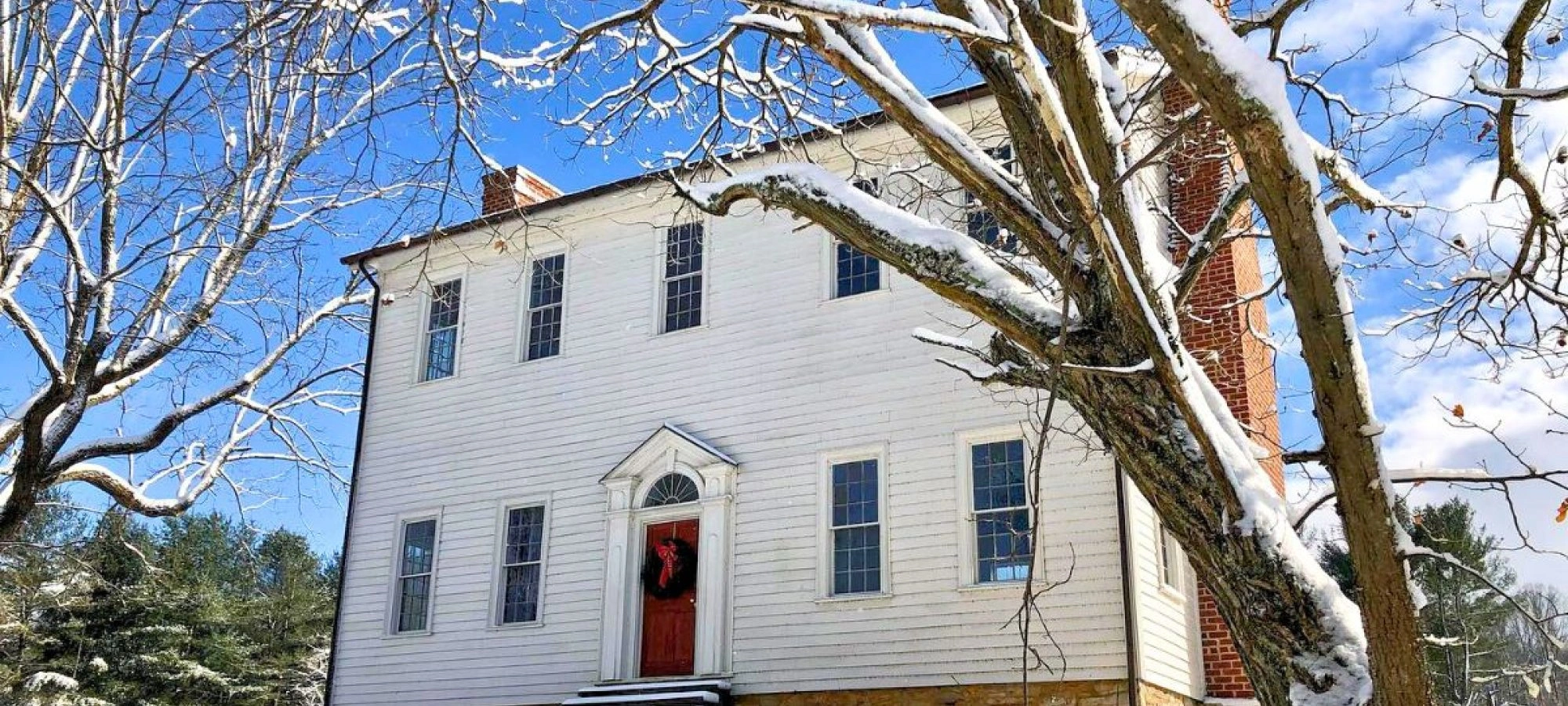 Historic white house with a red door and wreath, surrounded by snow-covered trees, under a bright blue sky. Wintery, peaceful atmosphere.
