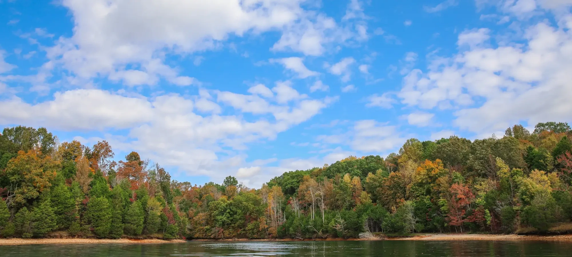 A serene autumn landscape with a calm lake reflecting the vibrant fall foliage of green, orange, and red trees under a bright blue, cloud-dotted sky.