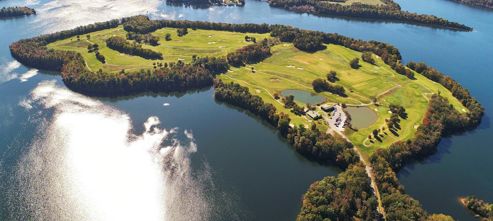 Aerial view of a lush golf course on an island surrounded by a shimmering lake. The landscape features vibrant greens, trees, and small ponds under a clear sky.