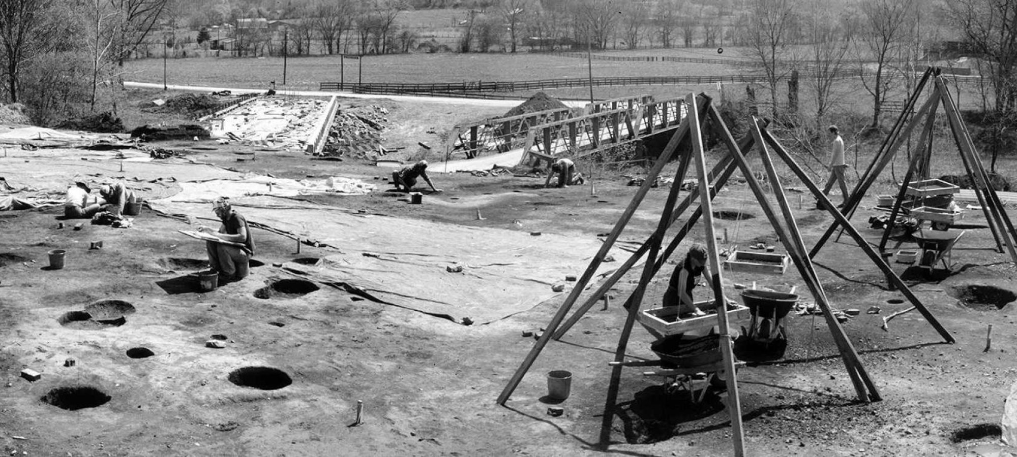 Archaeological dig site in a rural area, featuring workers examining the ground and using sieve stands. Large holes and tools are visible, with trees and fields in the background.