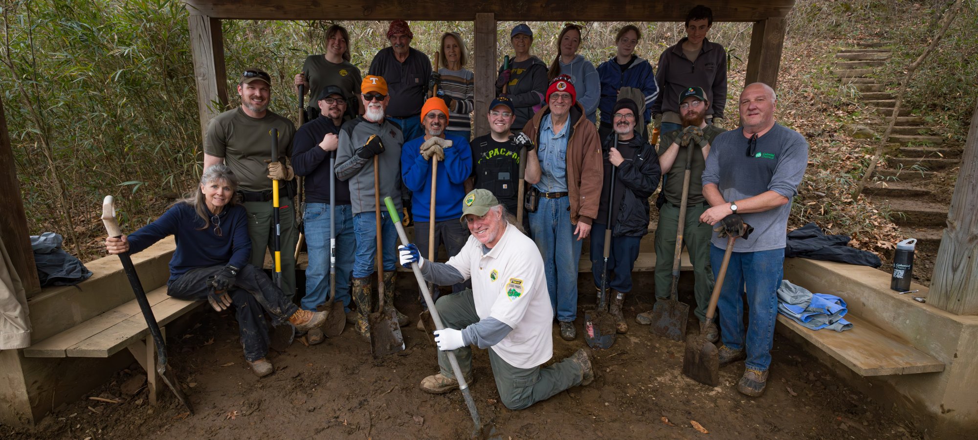 A group of 16 people, smiling and holding tools, poses under a wooden shelter in a wooded area. The mood is upbeat and collaborative.
