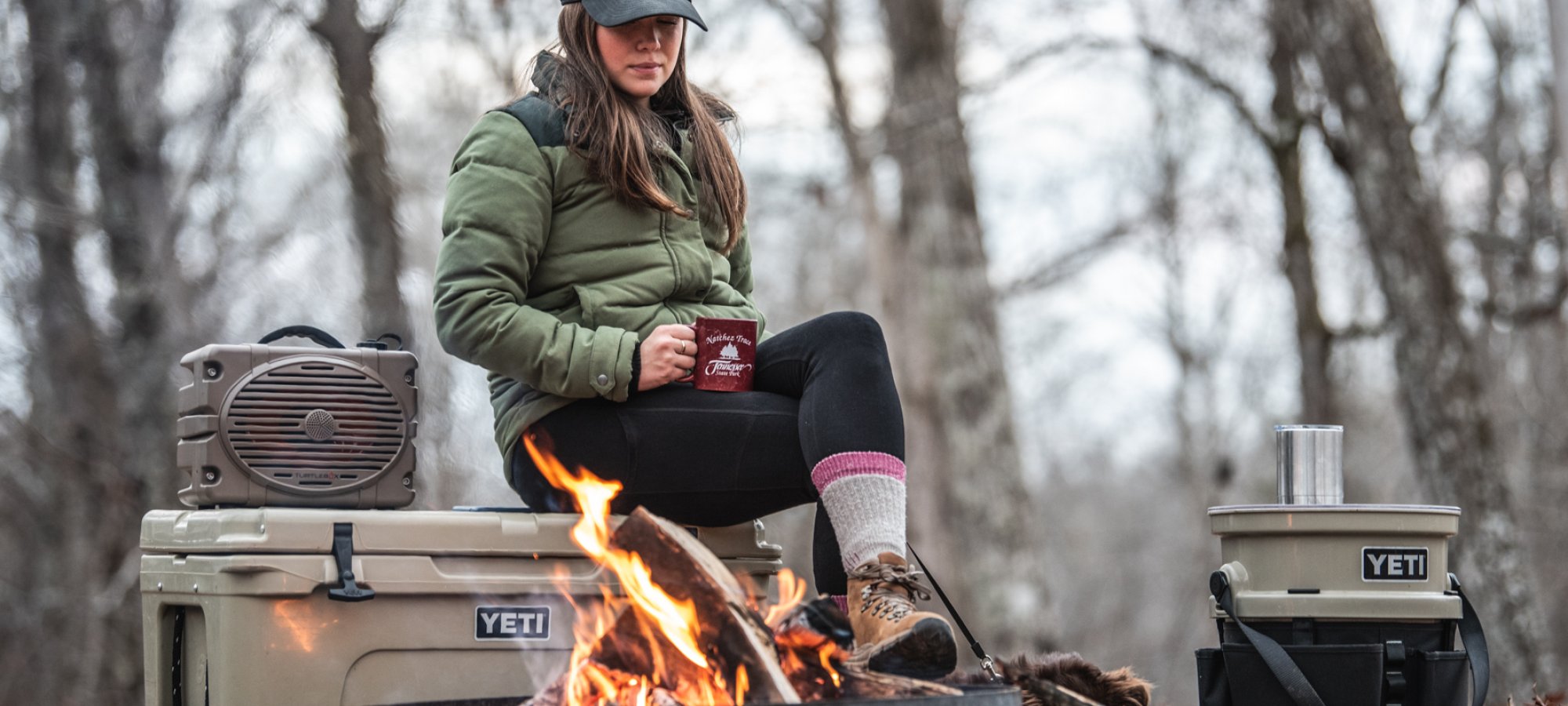 Woman in winter attire sits on a YETI cooler by a campfire in a forest, holding a mug. She looks relaxed, surrounded by camping gear.