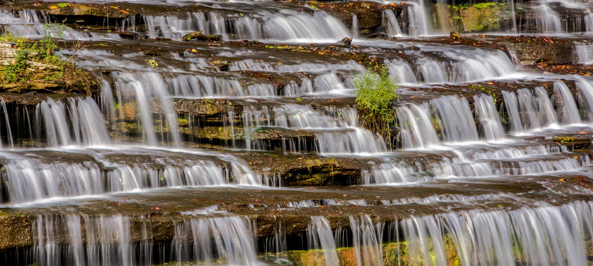 Tiered waterfall cascades down layered rock formations, surrounded by greenery. 