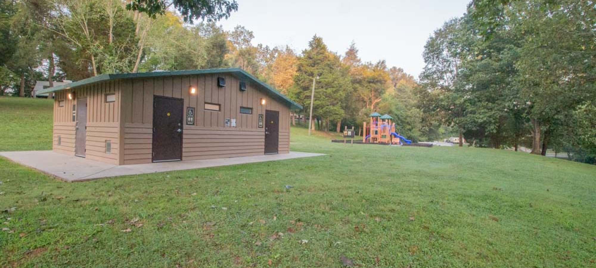 A small brown park building with dark doors sits on a grassy field surrounded by trees. In the background, a colorful playground is visible, evoking a serene, inviting atmosphere.