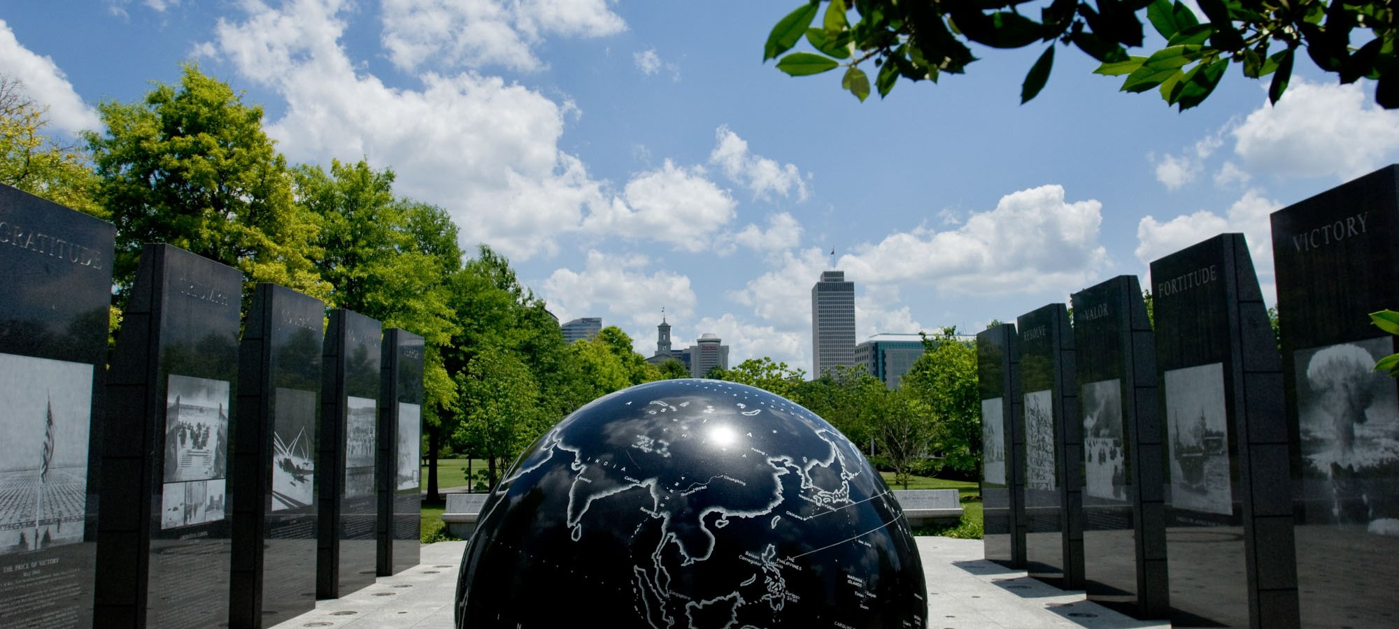 Black globe sculpture surrounded by tall engraved panels and trees under a blue sky.