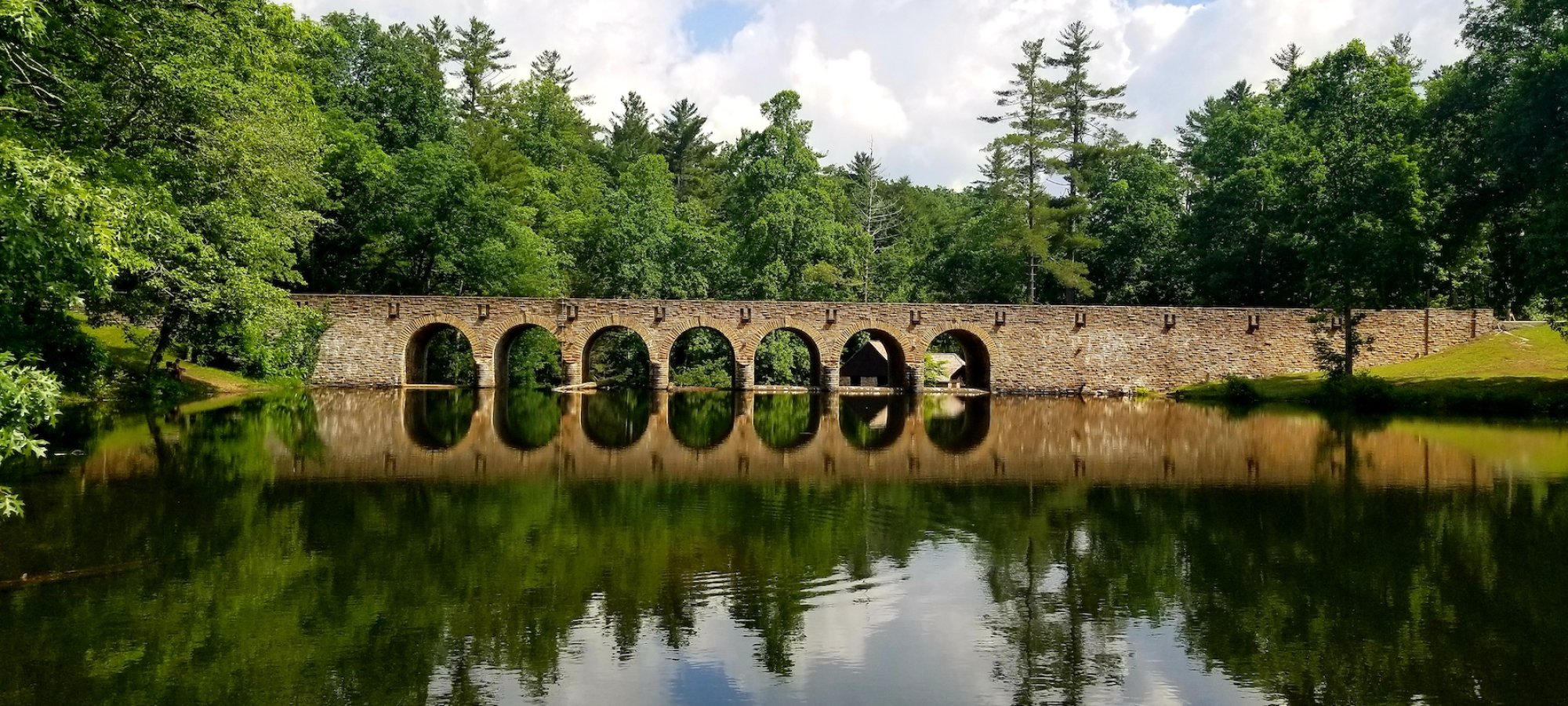 A stone bridge with multiple arches spans a calm river, reflecting a blue sky with fluffy clouds. Surrounded by lush green trees, the scene is serene.