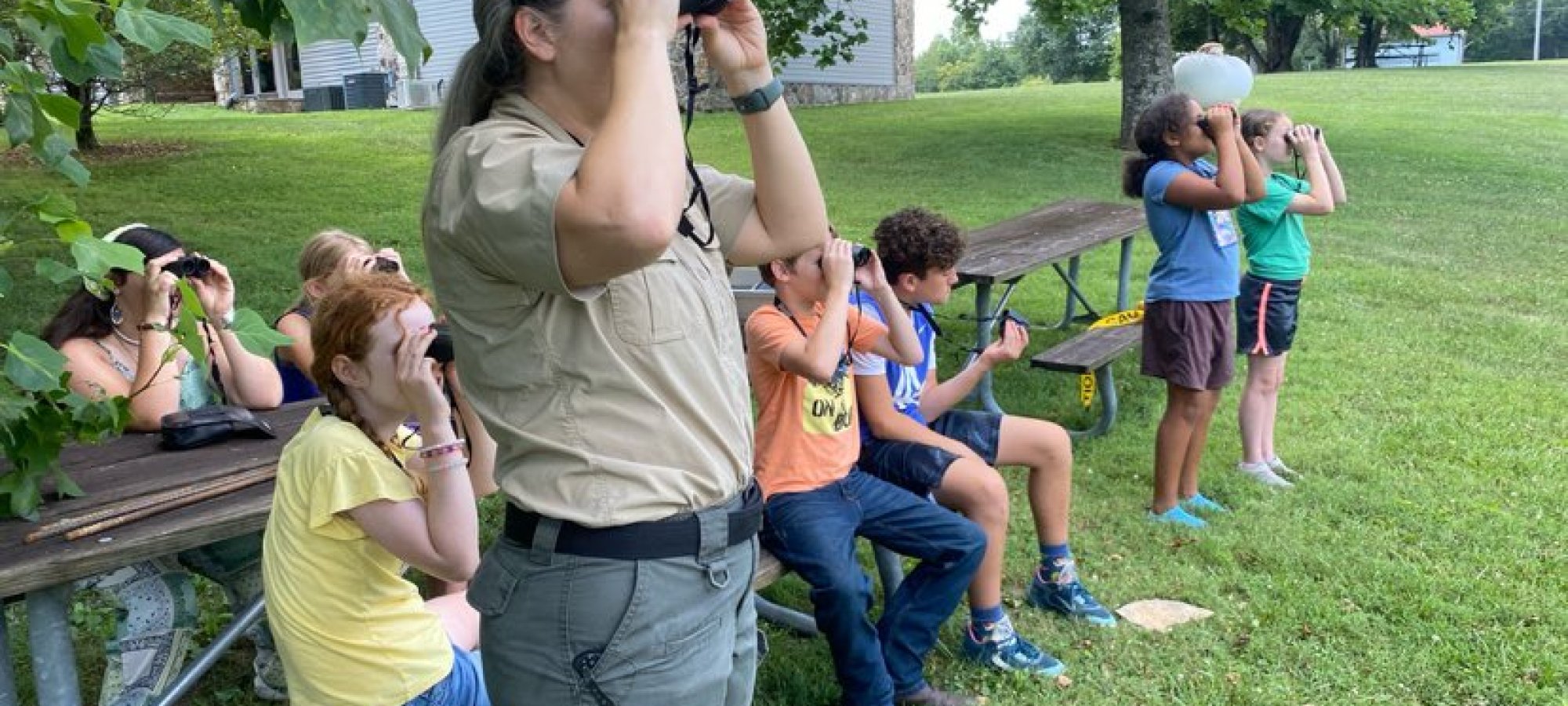 A group of children and a woman are birdwatching in a park. They sit and stand on grass near picnic tables, all looking through binoculars. It's a sunny day.