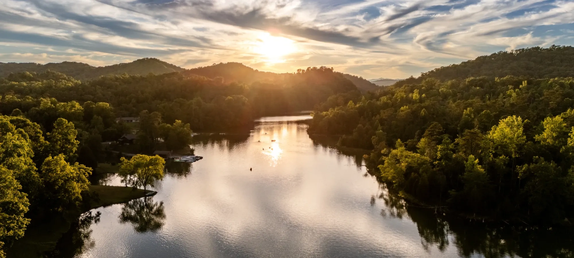 Sunset over a tranquil lake surrounded by lush green hills and dramatic clouds.