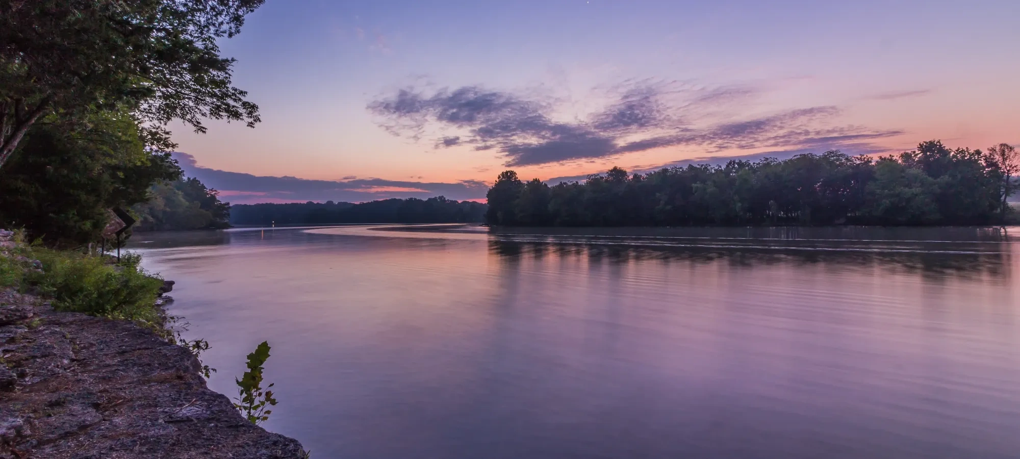 Calm river at sunset with trees and a purple sky.