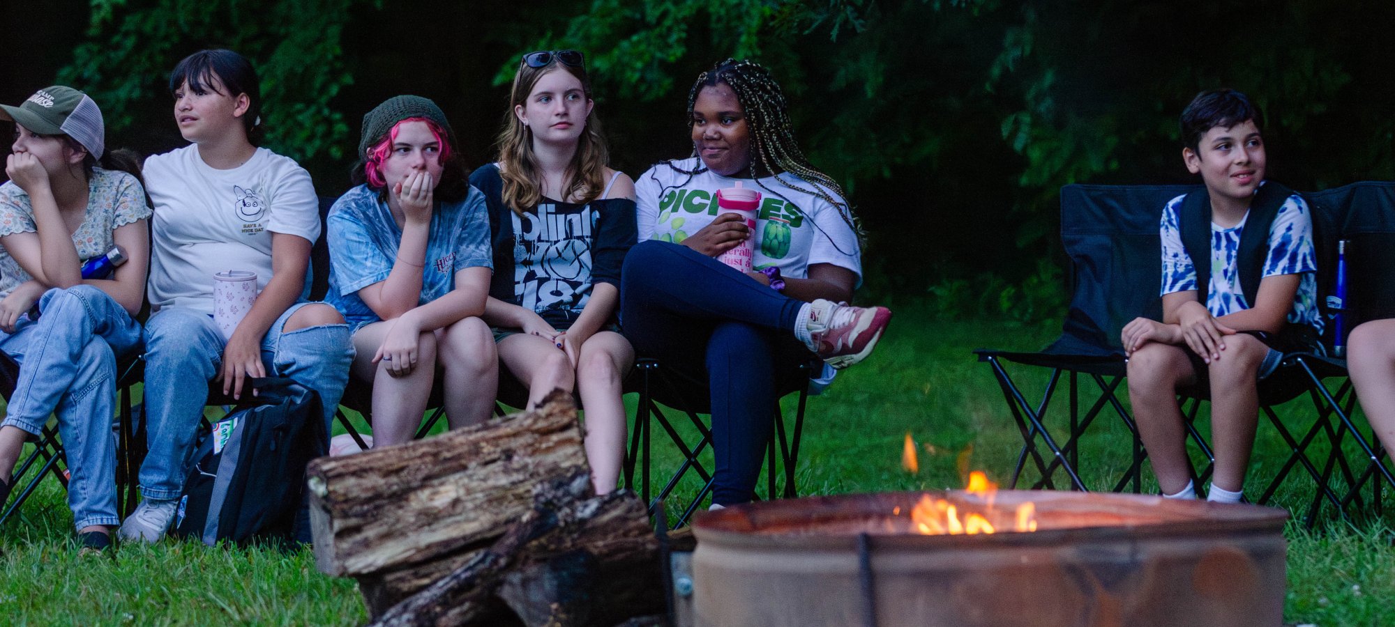 A group of young people sit around a campfire in chairs, with logs stacked nearby. They appear relaxed and engaged, surrounded by lush greenery.
