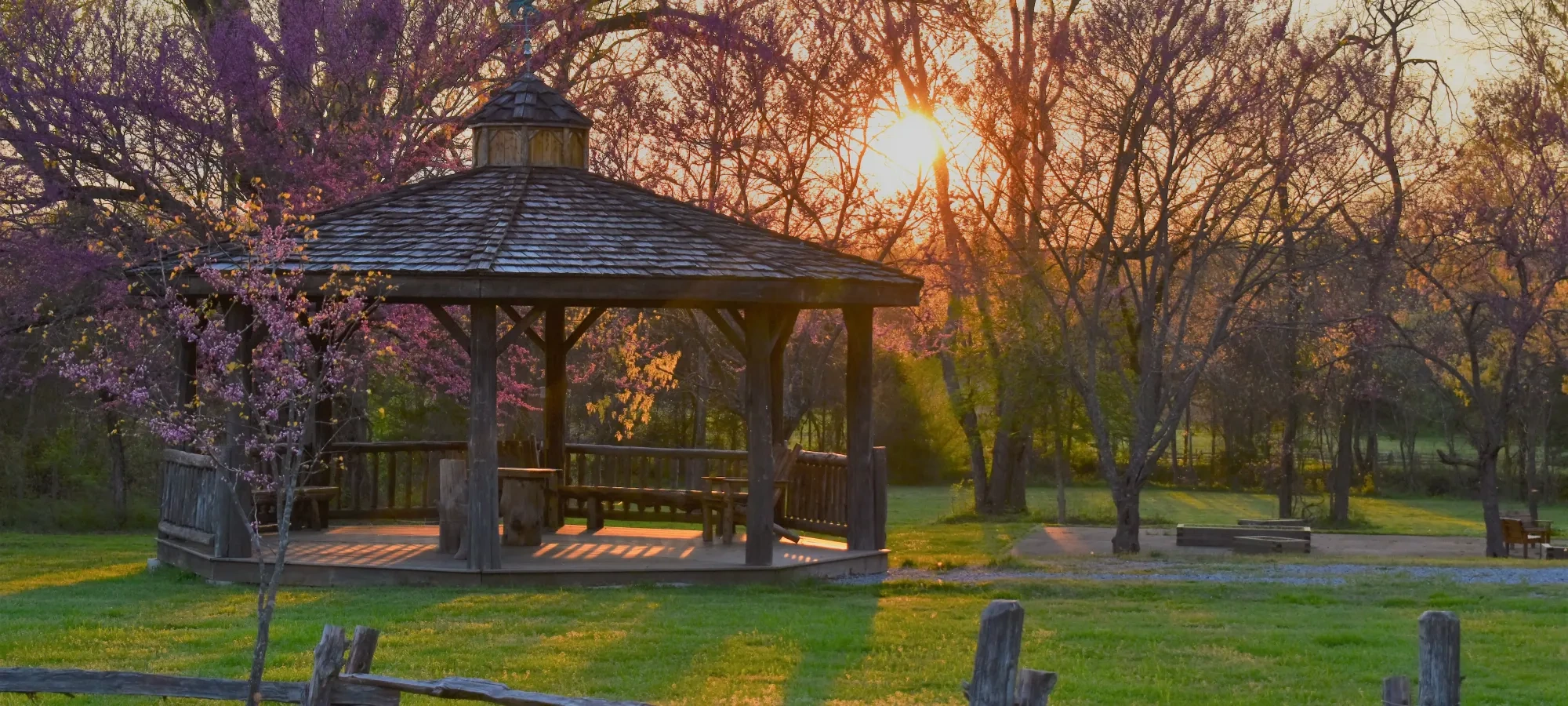 Sunset behind a wooden gazebo in a park with blooming trees.