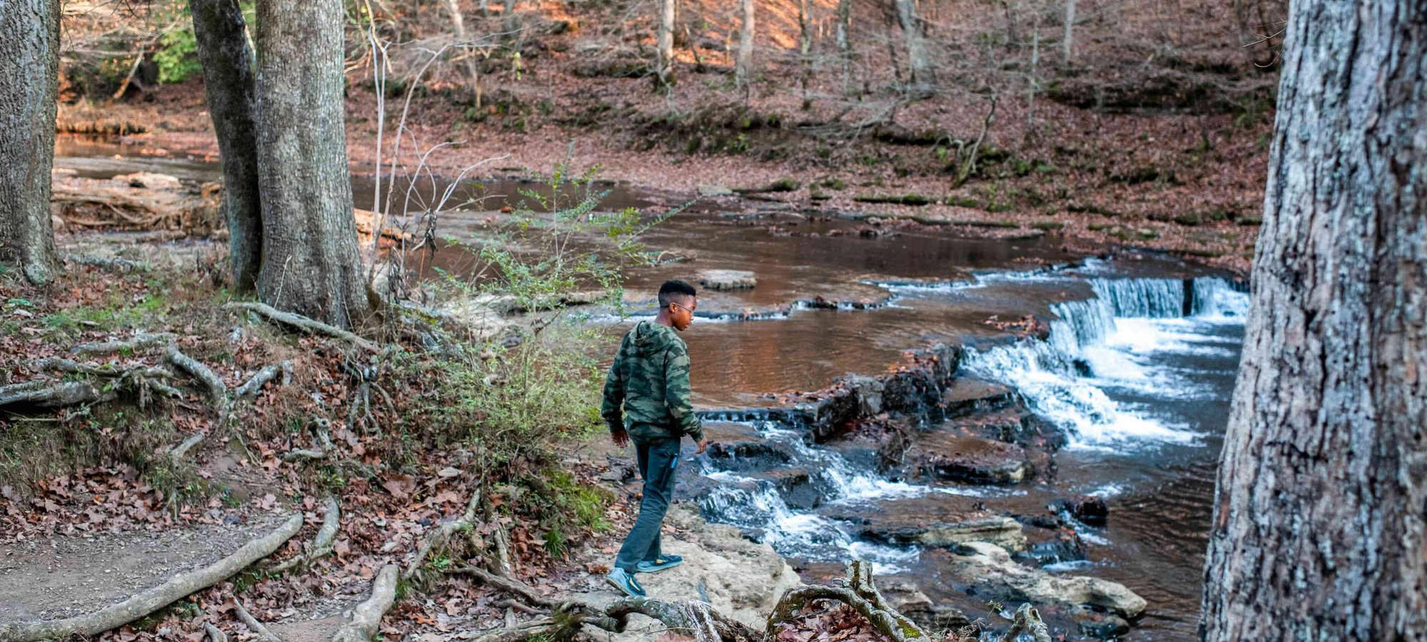 A person in a green jacket stands on rocks beside a small cascading waterfall in a forest. Tree trunks and fallen leaves surround the serene scene.
