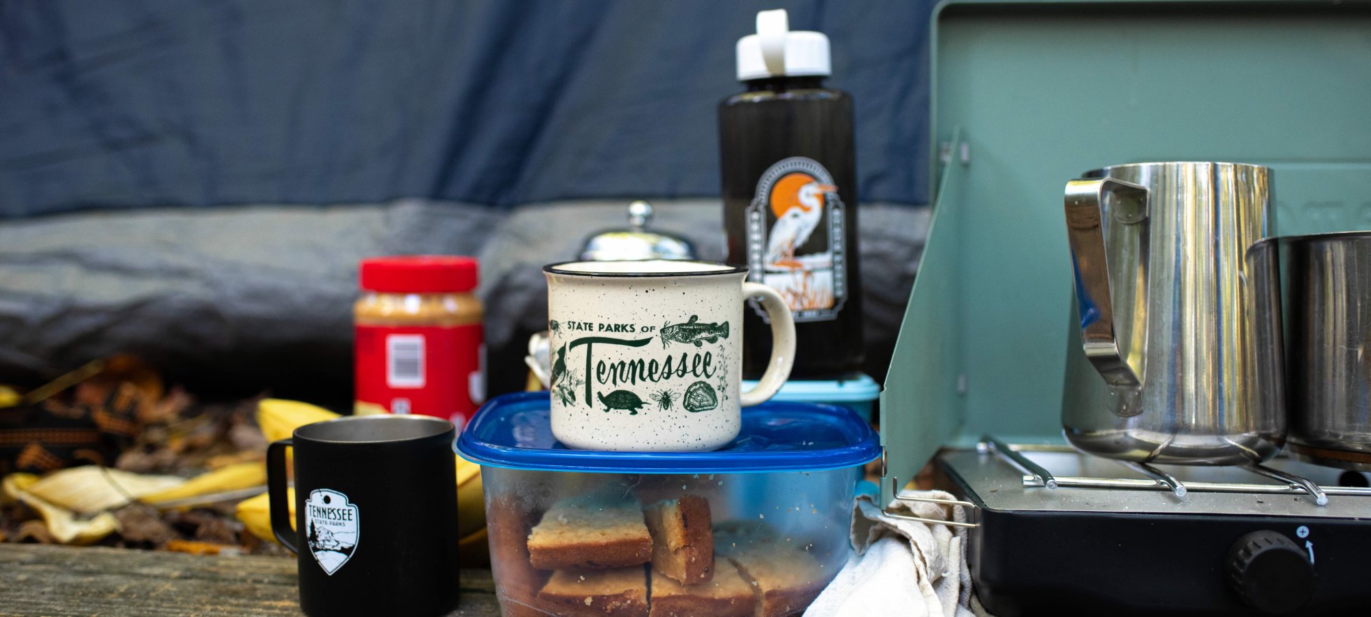 Camping scene with mugs, a container of sandwiches, peanut butter, water bottle, and kettle on a portable stove. Cozy and rustic outdoor vibe.