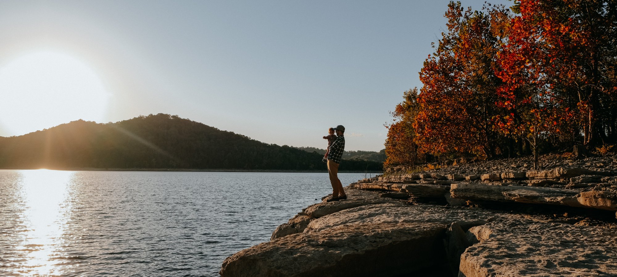 Person photographing a serene lake at sunset from a rocky shore, surrounded by autumn trees. Warm glow and peaceful ambiance fill the scene.