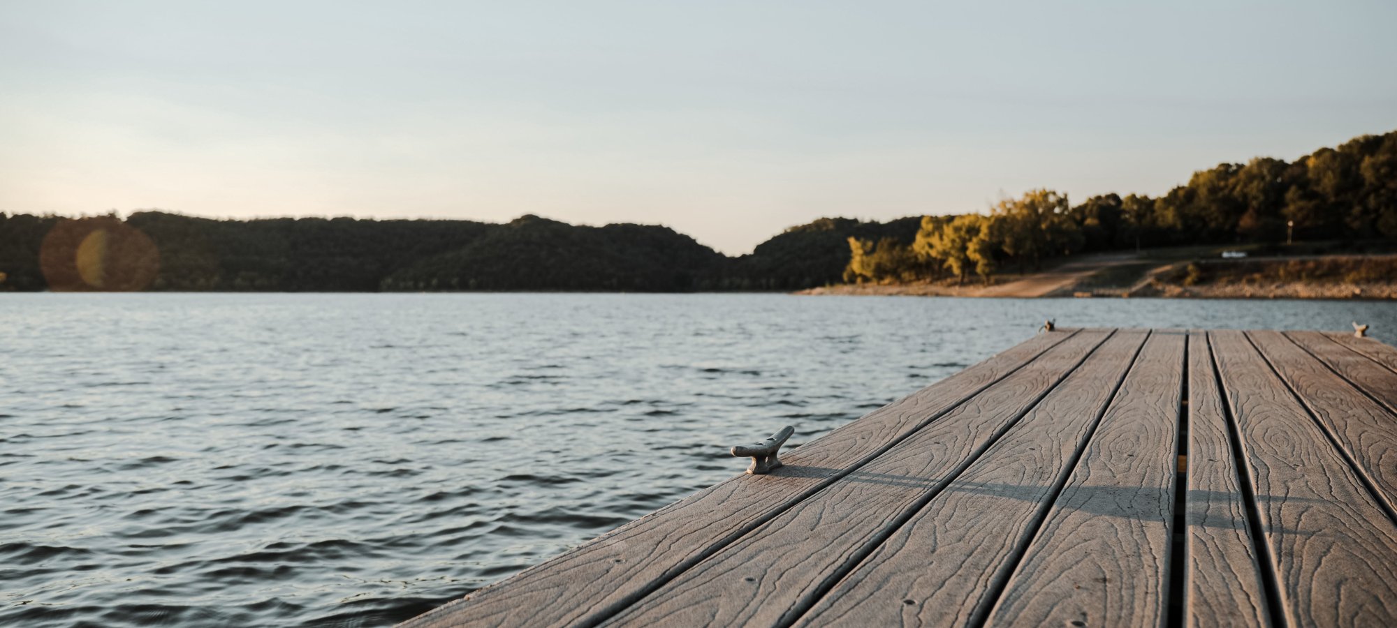 Wooden dock extending into a calm lake under a clear sky, surrounded by trees on distant hills, creating a peaceful, serene atmosphere at sunset.