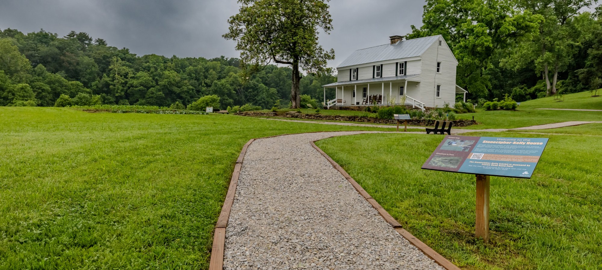 A gravel path leads to a white farmhouse surrounded by green grass and trees under a cloudy sky. A sign stands on the right, adding historical context.