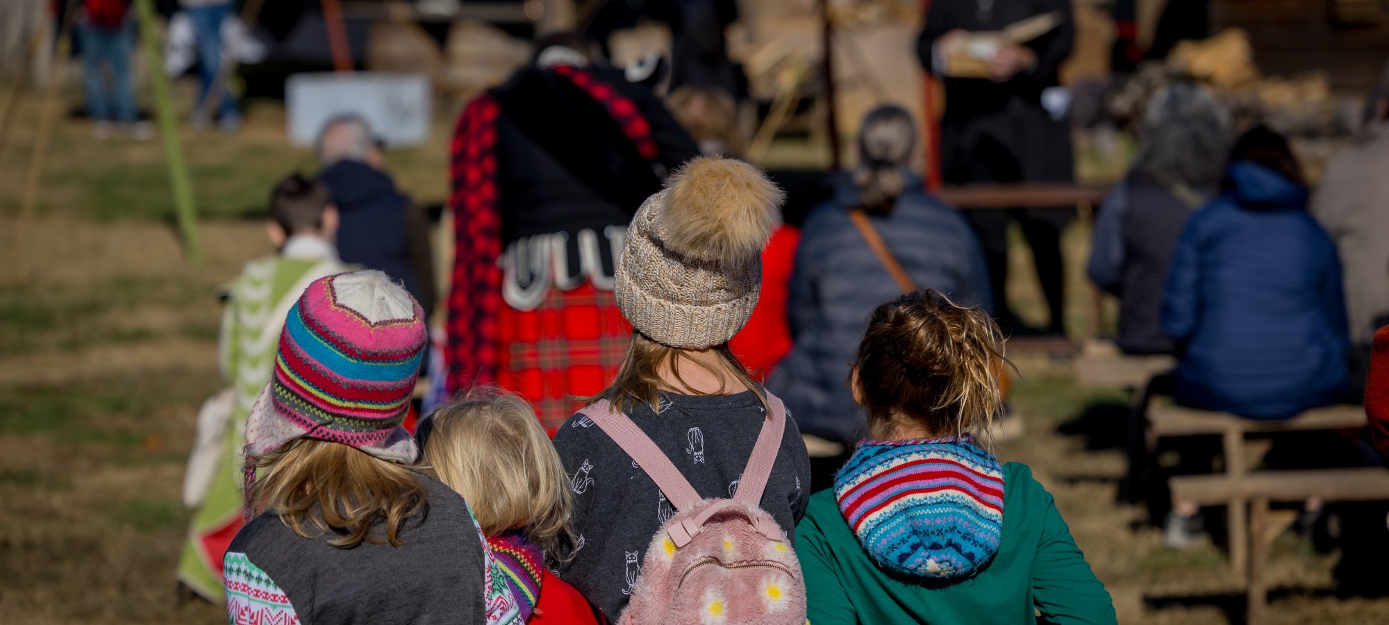 A group of children in colorful winter clothing sit on benches facing a person reading or speaking outdoors. The atmosphere is calm and attentive.