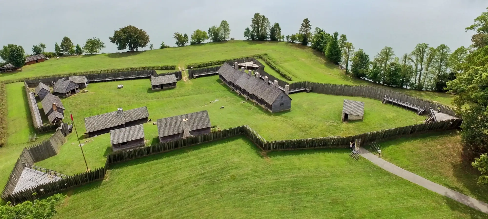 Aerial view of a historic fort with wooden buildings, surrounded by lush greenery and a lake.