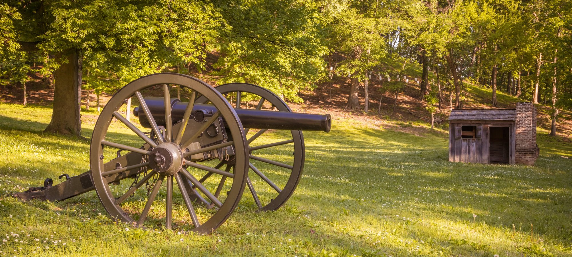 A historic cannon sits on a sunlit grassy field beside a small rustic wooden hut, with lush green trees in the background, evoking a serene, timeless setting.