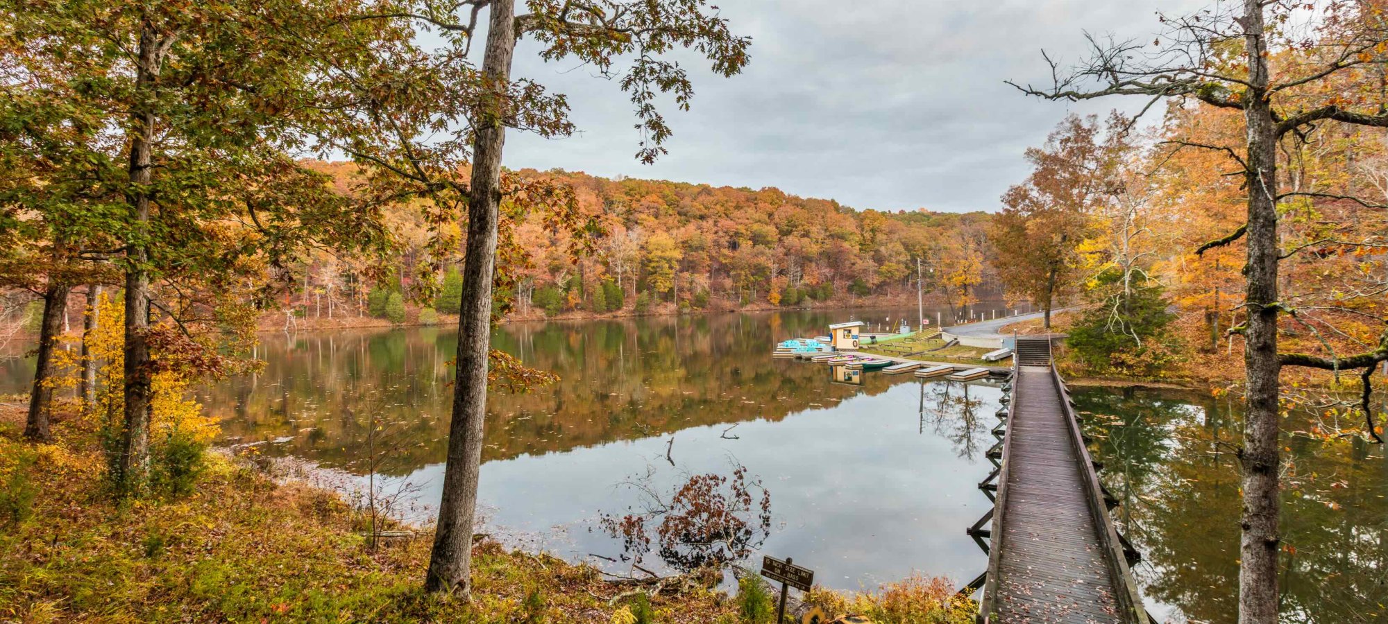 A serene autumn scene with a clear lake reflecting colorful fall trees. A narrow boardwalk extends into the water, surrounded by vibrant foliage.