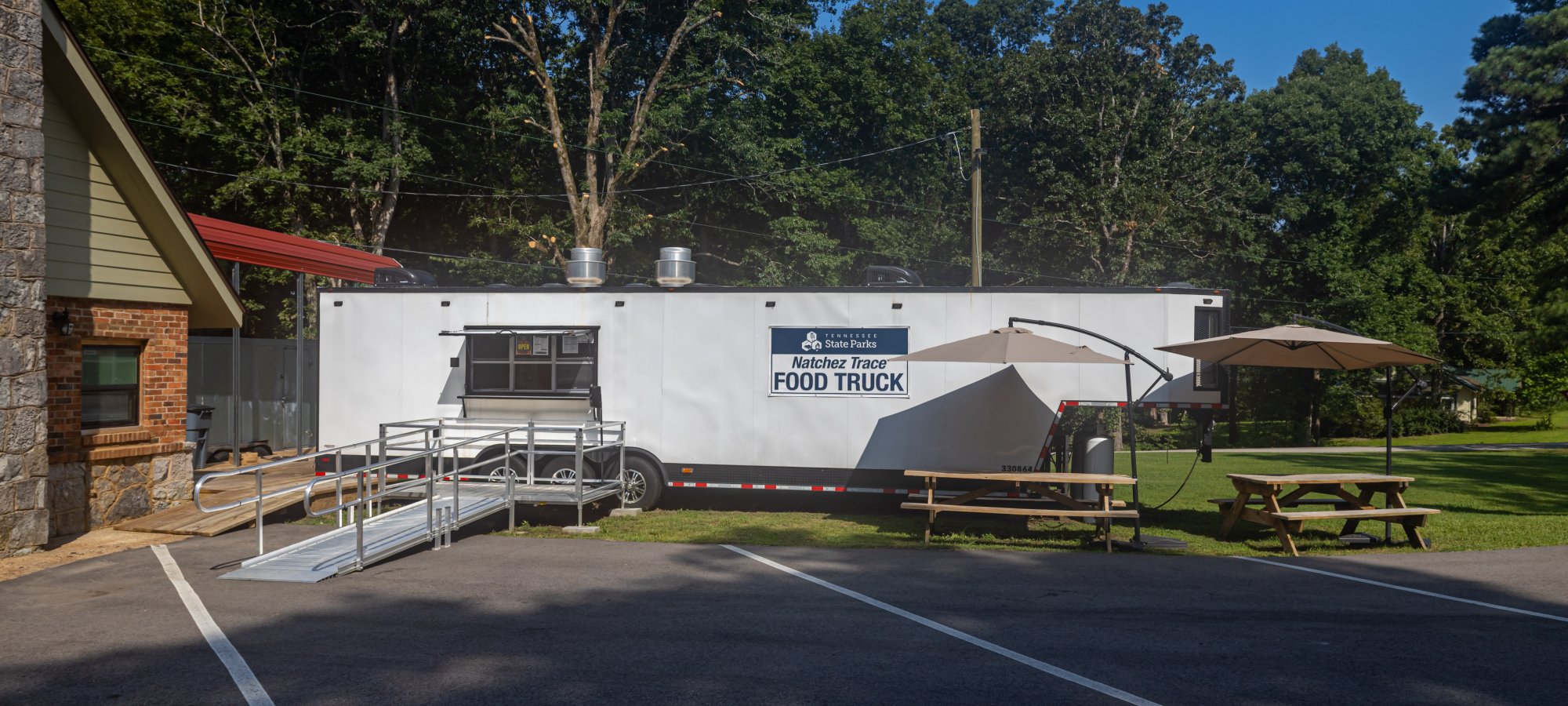 A white food truck in a park, shaded by tall trees and a clear blue sky. Nearby are picnic tables with umbrellas, creating a relaxed outdoor eating area.