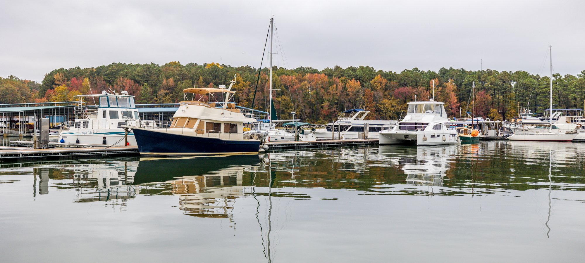 Boats are docked in a marina with calm water reflecting their image. The background shows autumn trees with orange, red, and green foliage under a cloudy sky.