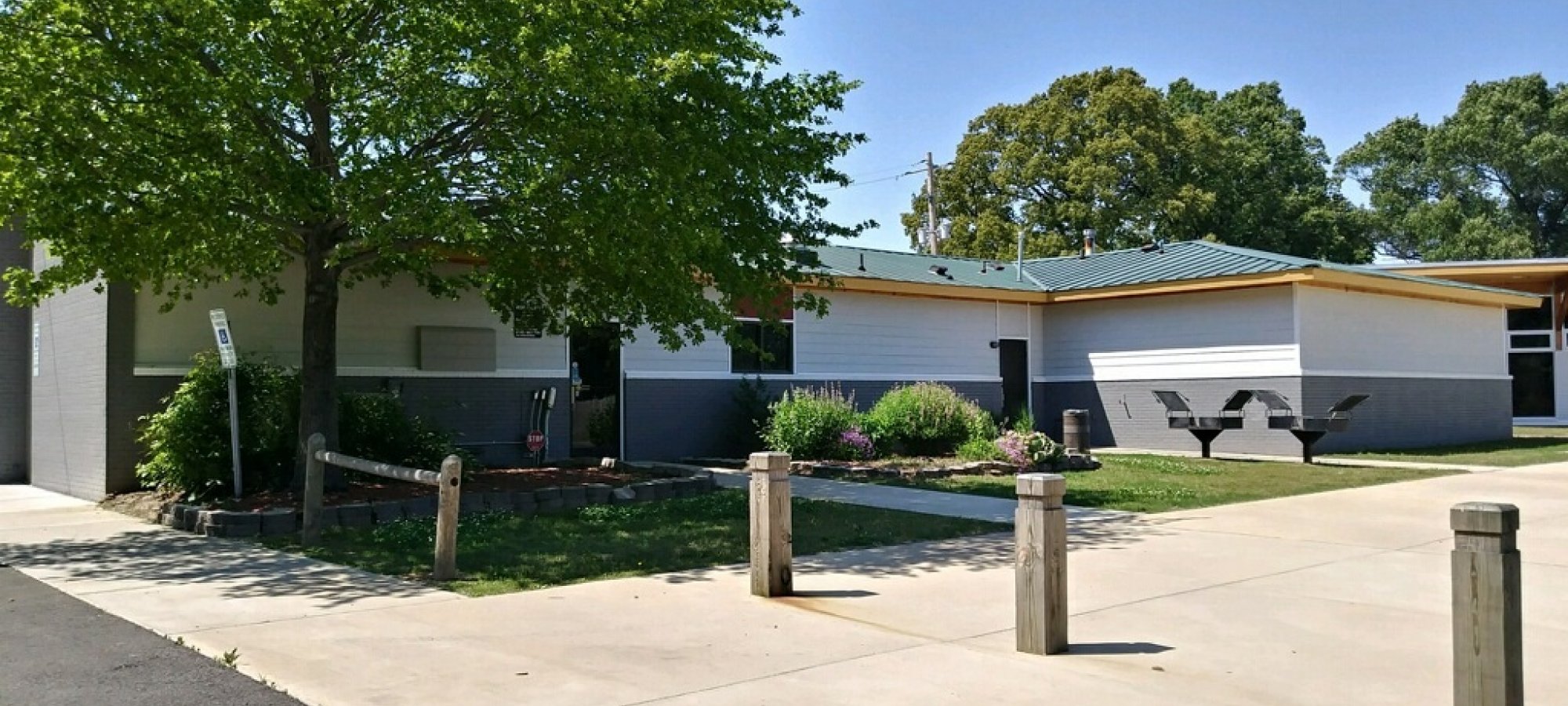 A modern, single-story building with a green roof sits surrounded by trees and bushes under a clear blue sky. A sidewalk and picnic table are nearby.