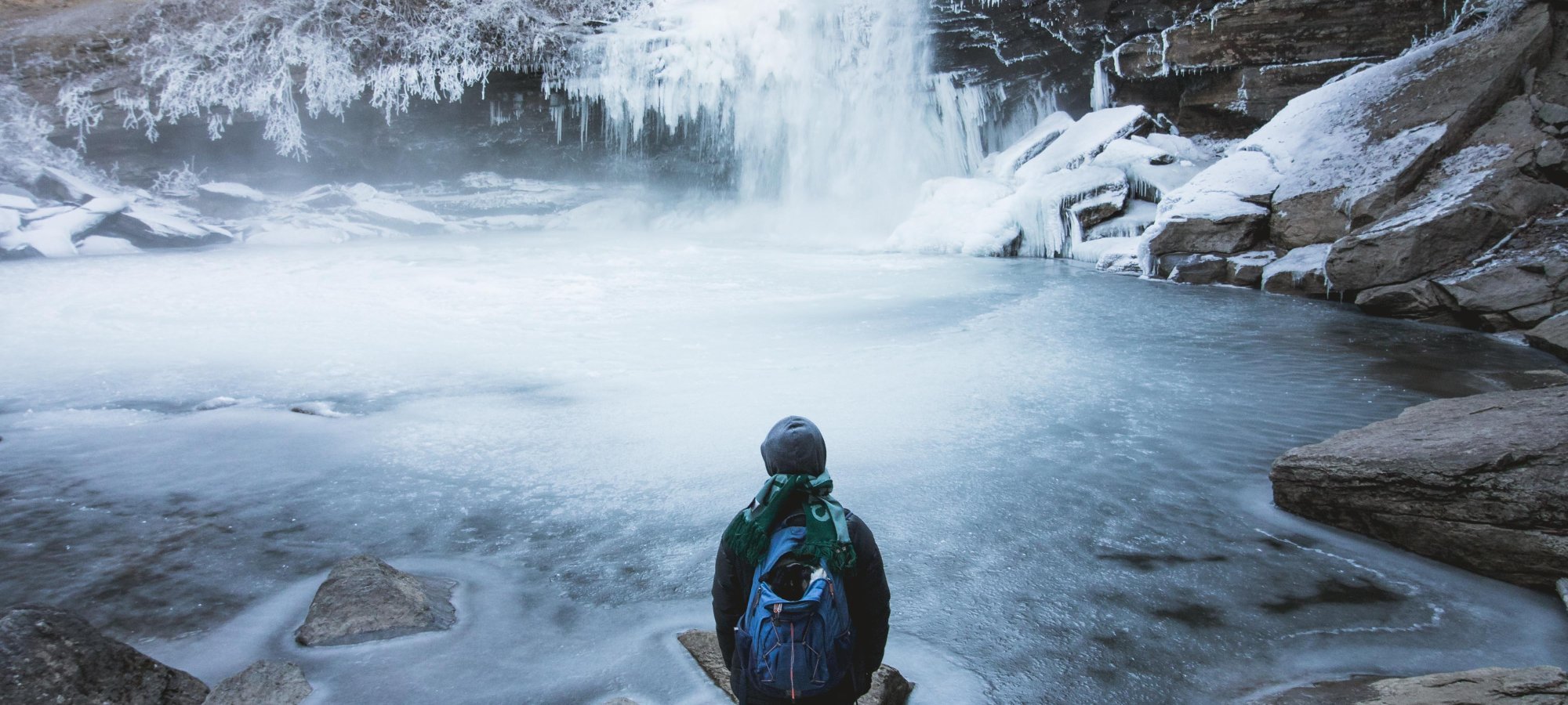 Person with a backpack stands on rocks, gazing at a frozen waterfall and icy lake, surrounded by frosted trees, evoking a serene, winter scene.