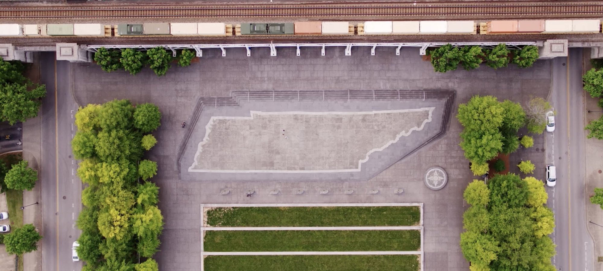 Aerial view of paved plaza featuring a large, outlined Tennessee shape. Train tracks run above, bordered by lush green trees lining the pathways.