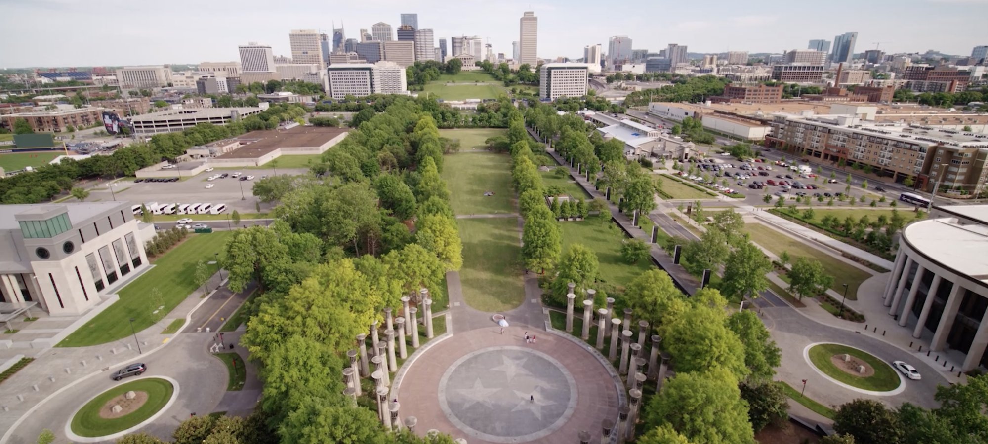 Aerial view of a city park with a circular plaza featuring a star pattern. Tree-lined paths extend towards a city skyline under a clear blue sky.