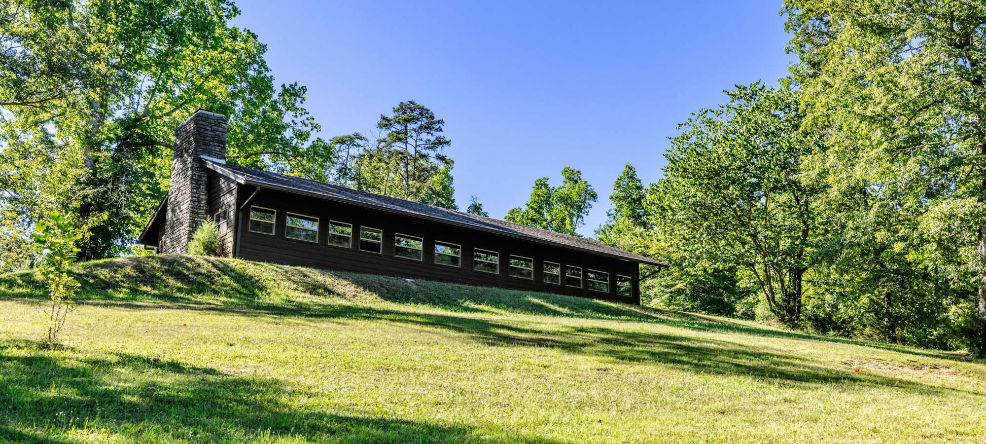 A long, dark brown house with large windows sits on a sunlit hillside, surrounded by lush green trees under a clear blue sky, evoking tranquility.