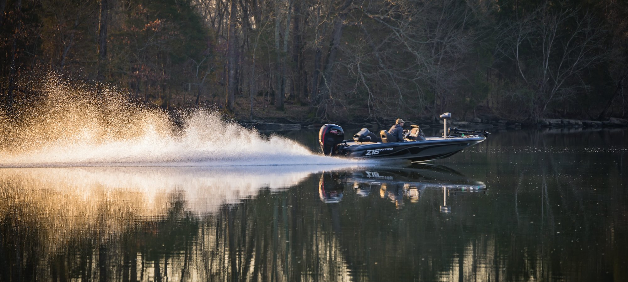 A speedboat creates a trail of white spray as it skims across a calm lake, surrounded by bare trees, reflecting a serene winter scene at dusk.