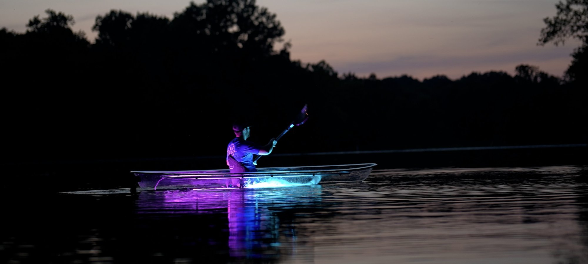 A person kayaks on a calm lake at dusk in a transparent kayak illuminated with vibrant blue and pink lights, creating a serene and magical atmosphere.