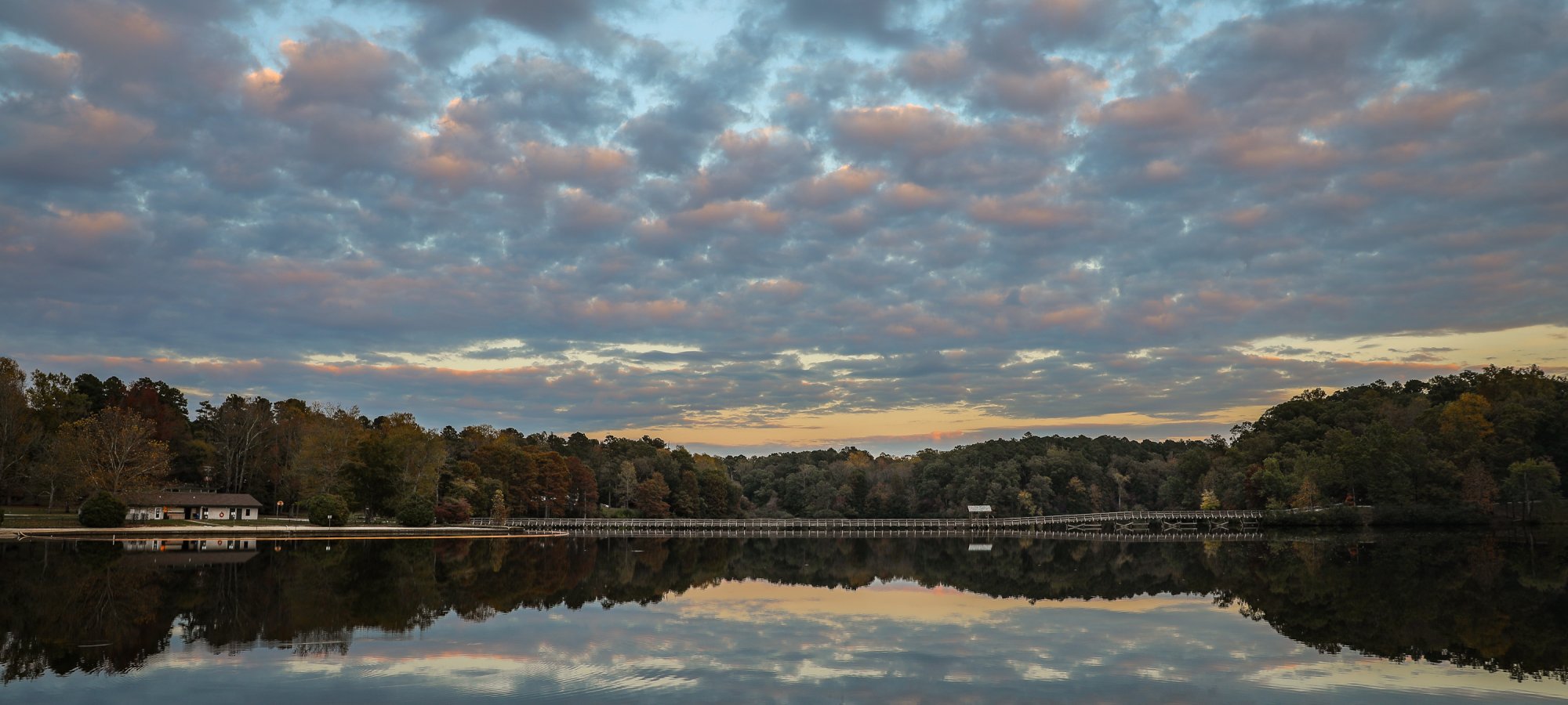 Calm lake reflecting a cloudy, colorful sky during sunset. Surrounding trees in autumn hues line the shore, creating a peaceful, serene scene.