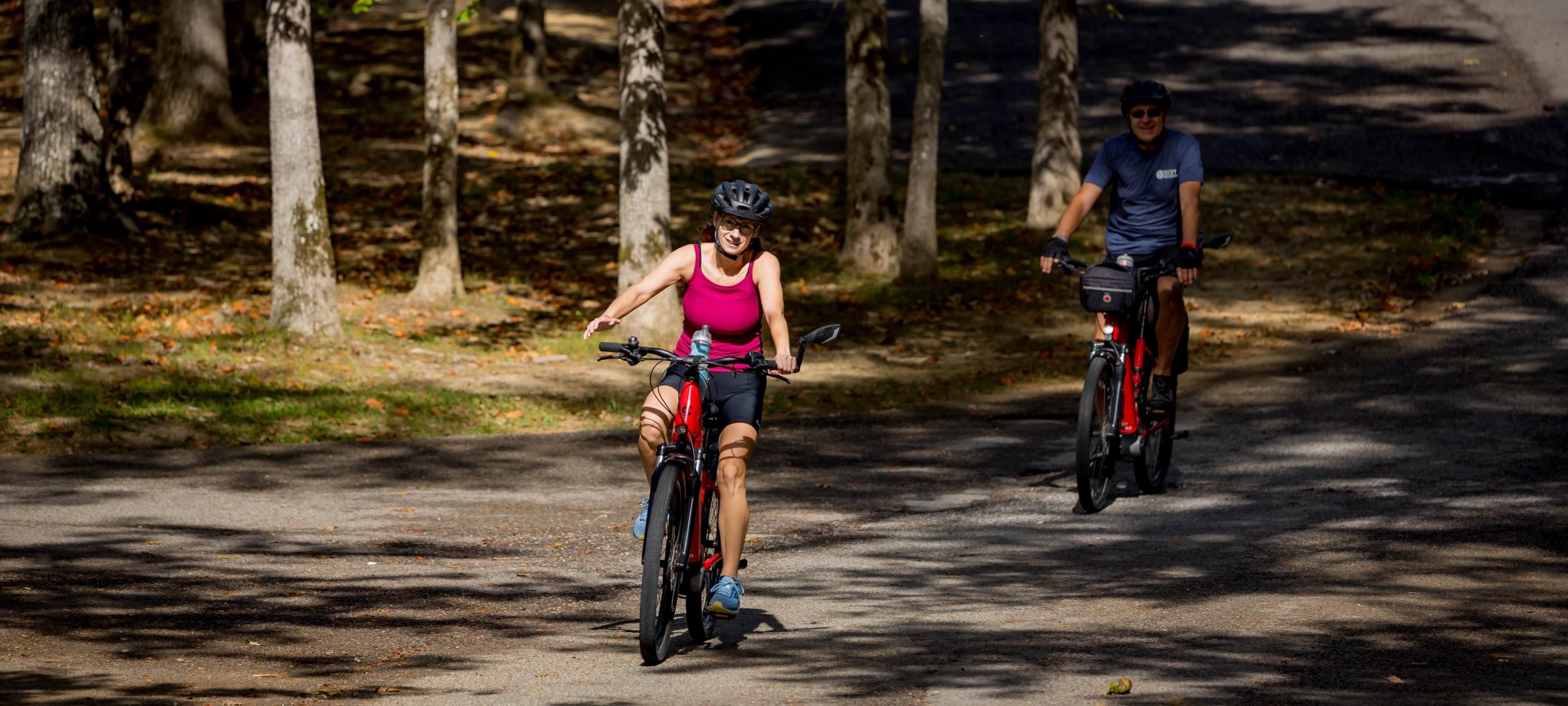 Two cyclists ride through a sunlit, tree-lined road. The leading cyclist wears a pink top and smiles, while the second cyclist follows, shaded by trees.