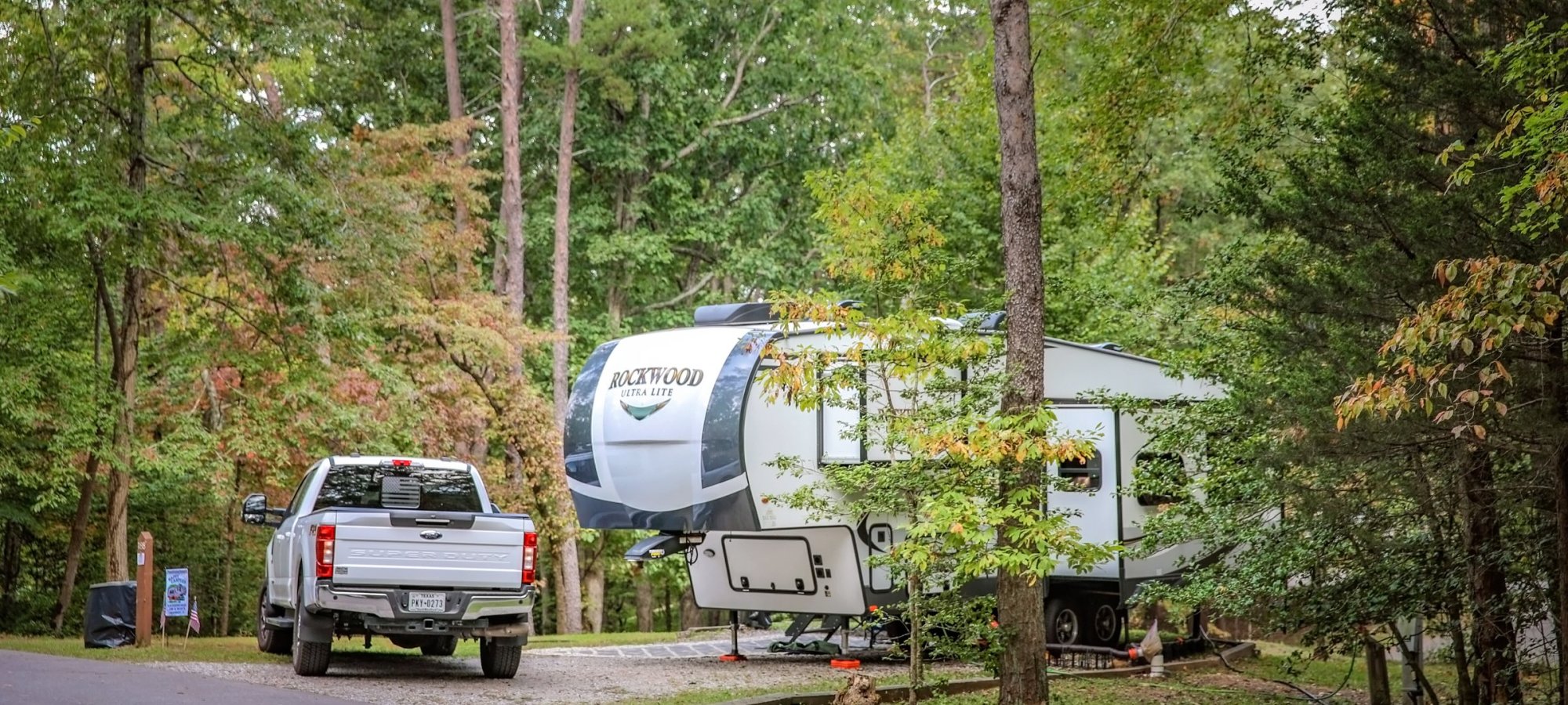 A white truck is parked next to a large RV surrounded by lush green trees in a serene forest setting, conveying a peaceful camping vibe.