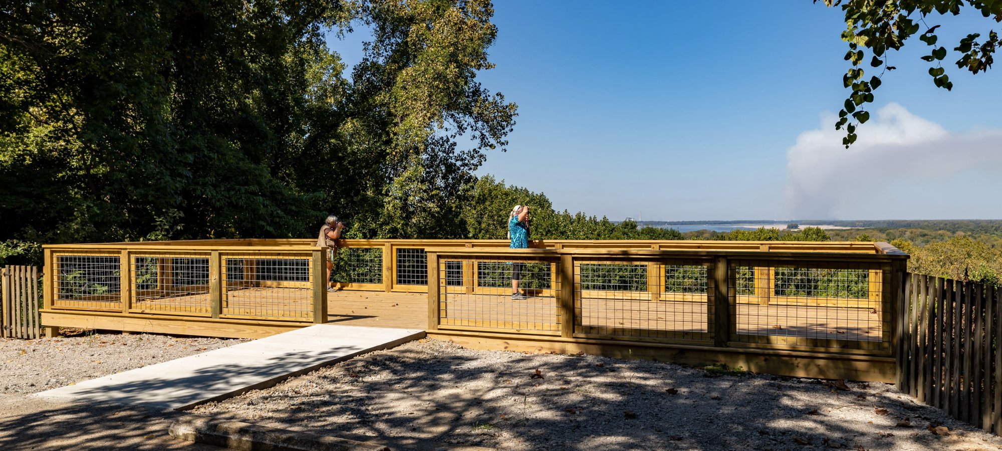 A wooden observation deck overlooks a forest with two people enjoying the view. The sky is clear blue with a distant smoke cloud on the horizon.