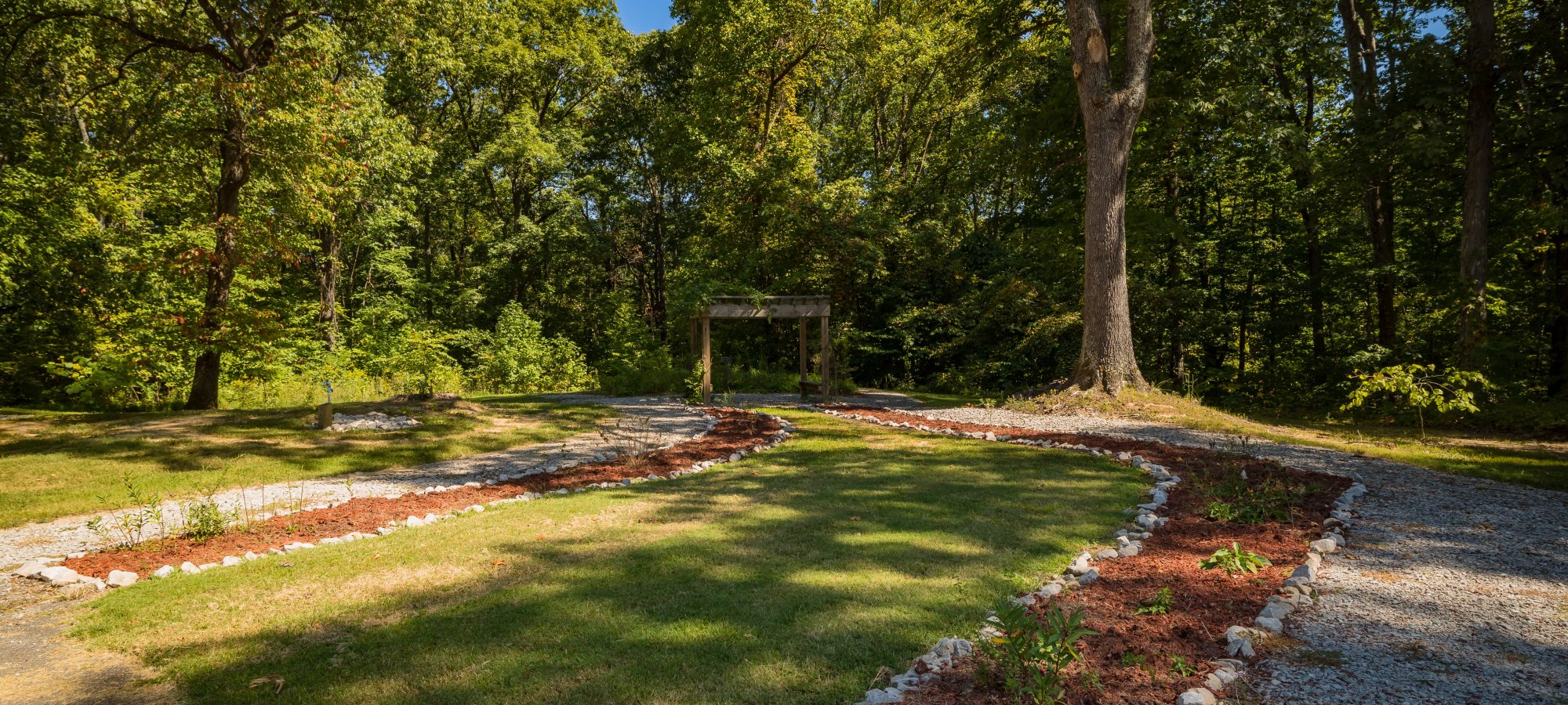A serene garden scene with a curved gravel path lined by small rocks and mulch, surrounded by lush green trees under a clear blue sky.