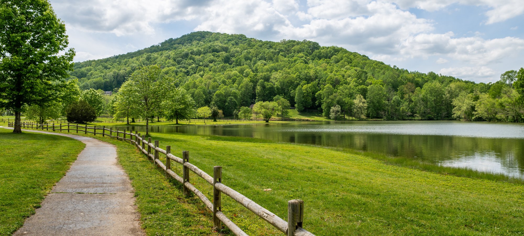 A winding path with a wooden fence leads to a lush green hill beside a tranquil lake under a partly cloudy sky, evoking a peaceful, serene atmosphere.