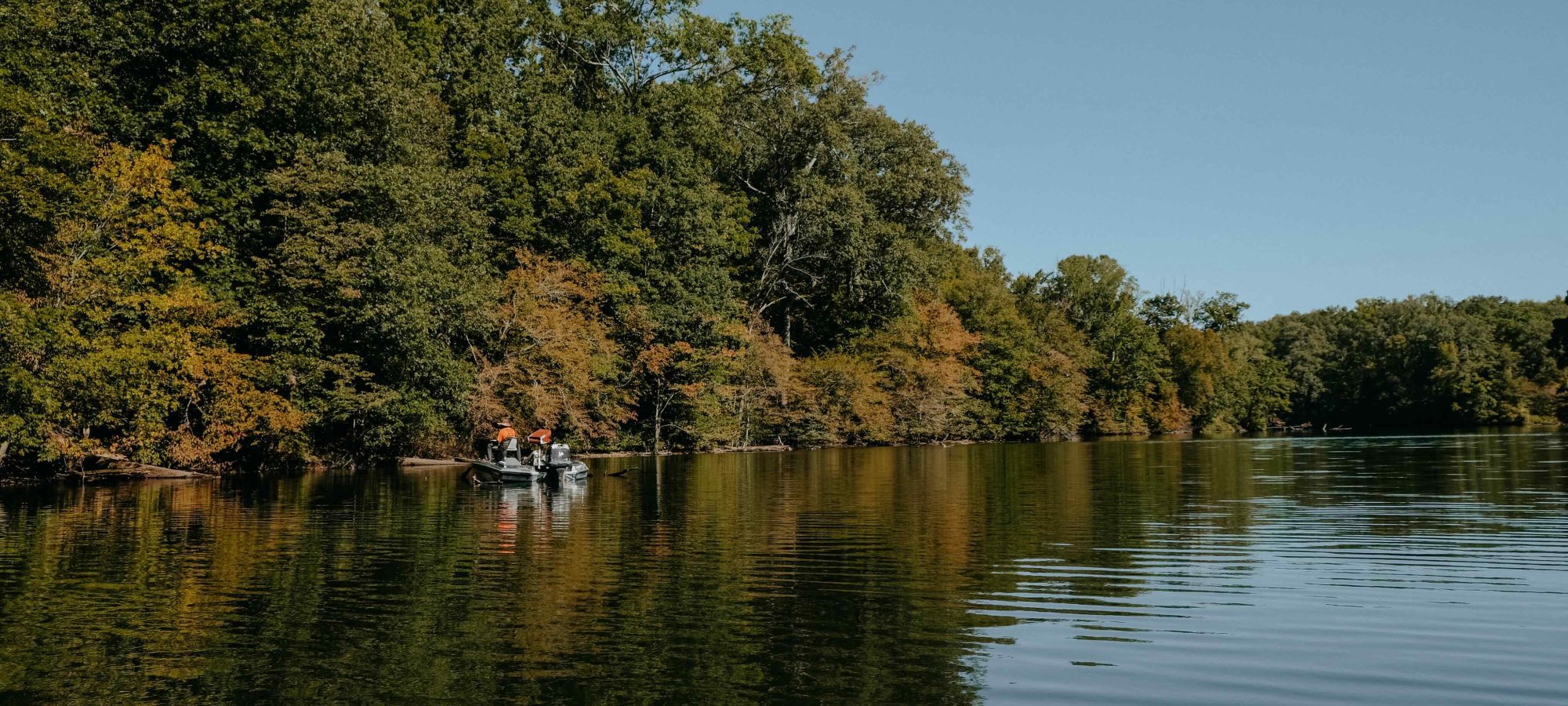A small boat floats on a calm lake, surrounded by lush green and slightly autumnal trees under a clear blue sky, conveying tranquility and nature.