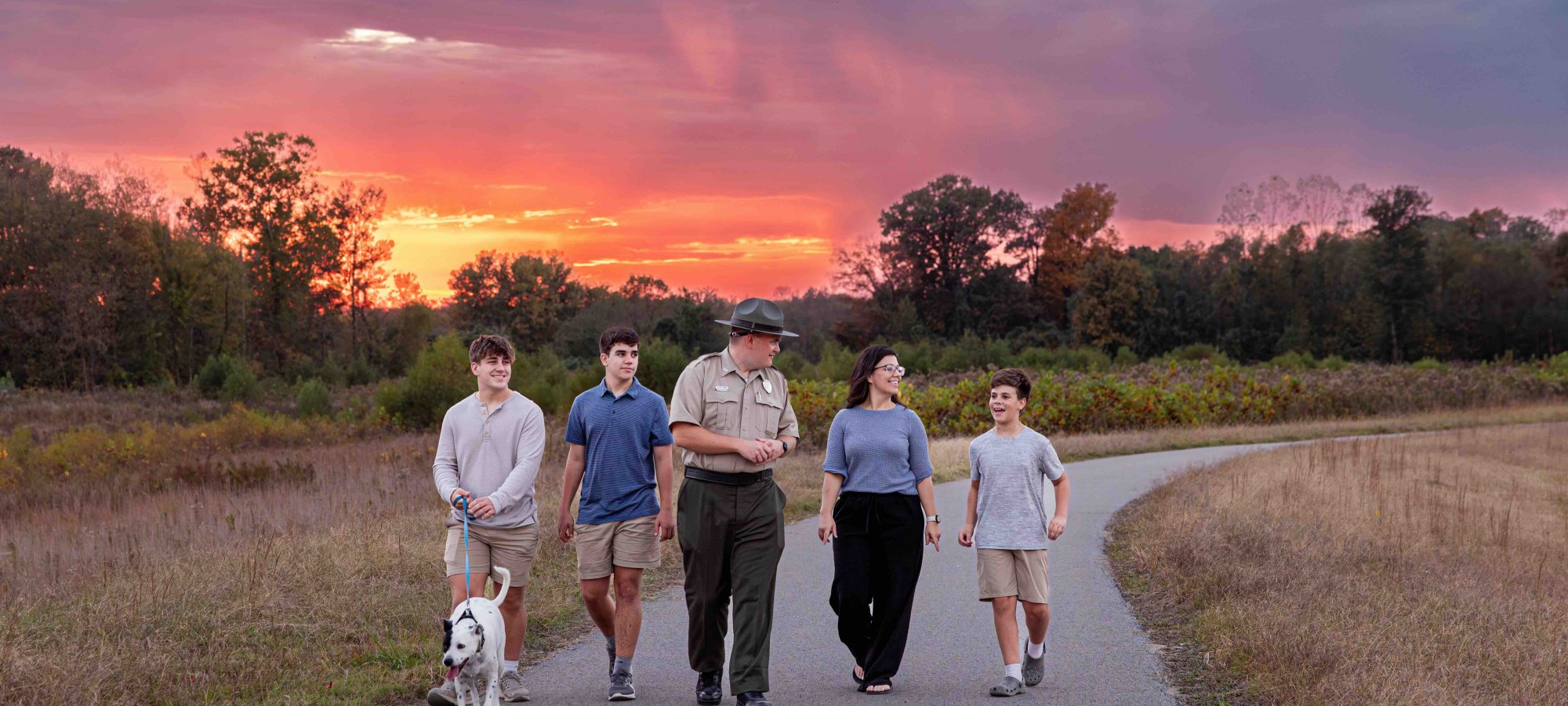 family with a dog and a ranger walking down a paved trail while the sun it setting