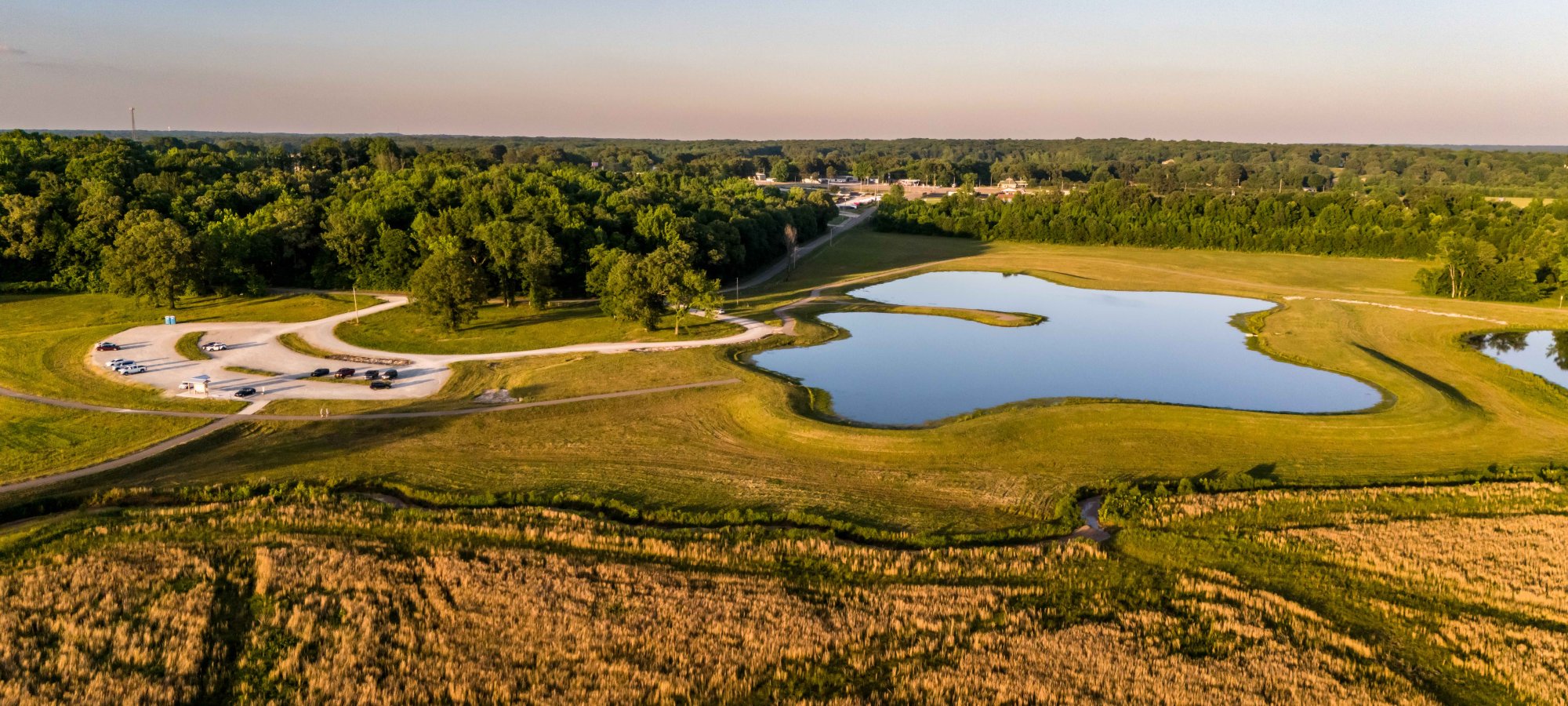 Aerial view of a tranquil park with a small lake, surrounded by green fields and dense forest. Cars are parked on a winding road. Sunny, peaceful setting.