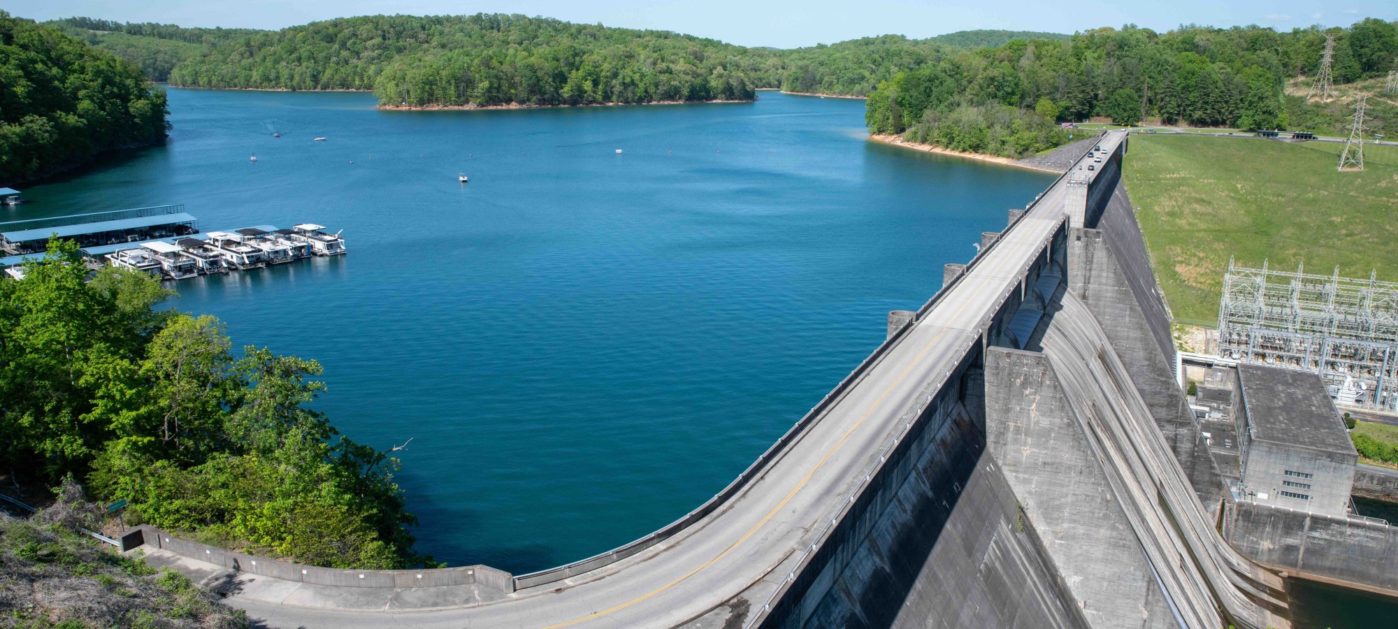 Aerial view of a large dam with a road above, surrounded by lush green forests and a calm blue lake. Boats docked to the left create a peaceful scene.