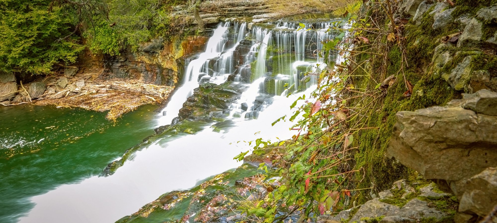 Cascading waterfall flows over tiered rocks into a serene green pool, surrounded by lush forest. A stone ledge with moss and leaves frames the scene.
