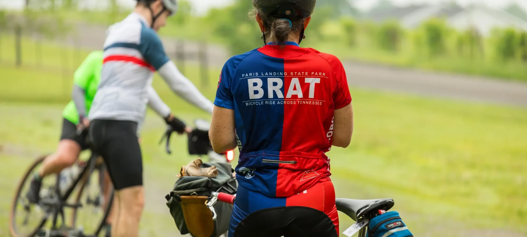 Cyclists in colorful jerseys resting with bicycles on a grassy path.