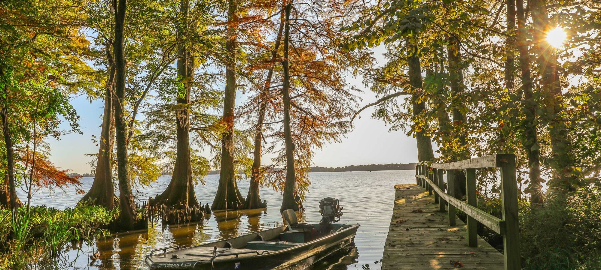 A serene lakeside scene with a motorboat moored beside a wooden dock. Tall trees with autumn foliage surround, and the sun shines warmly through the branches.