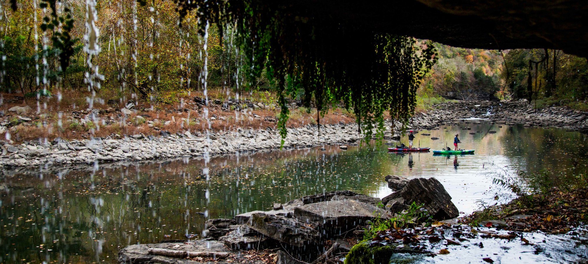 Serene river scene with kayakers paddling by rocky shoreline. Water drips from above, framing the view with greenery. Autumn foliage reflects in calm water.