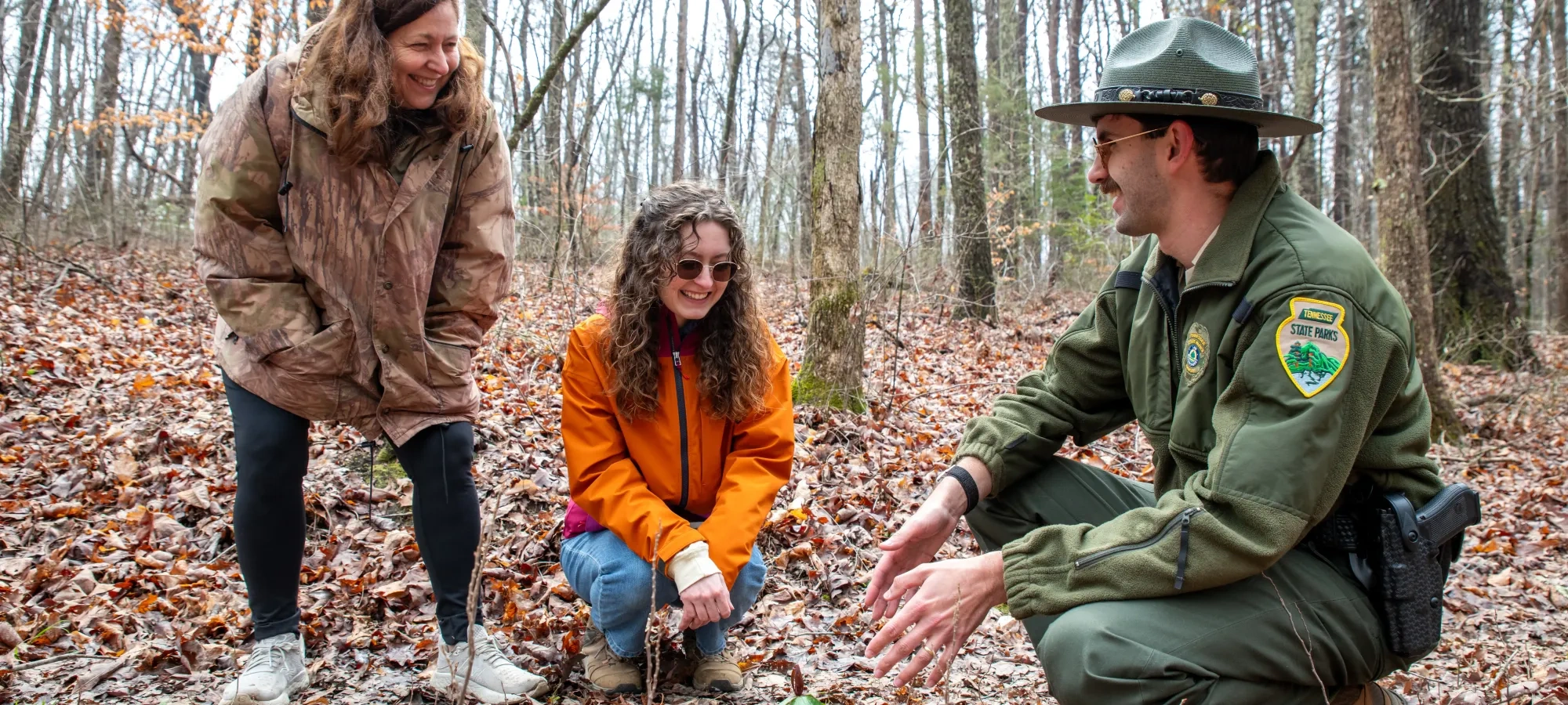 Park ranger and two women crouching in a forest, smiling and observing the ground.