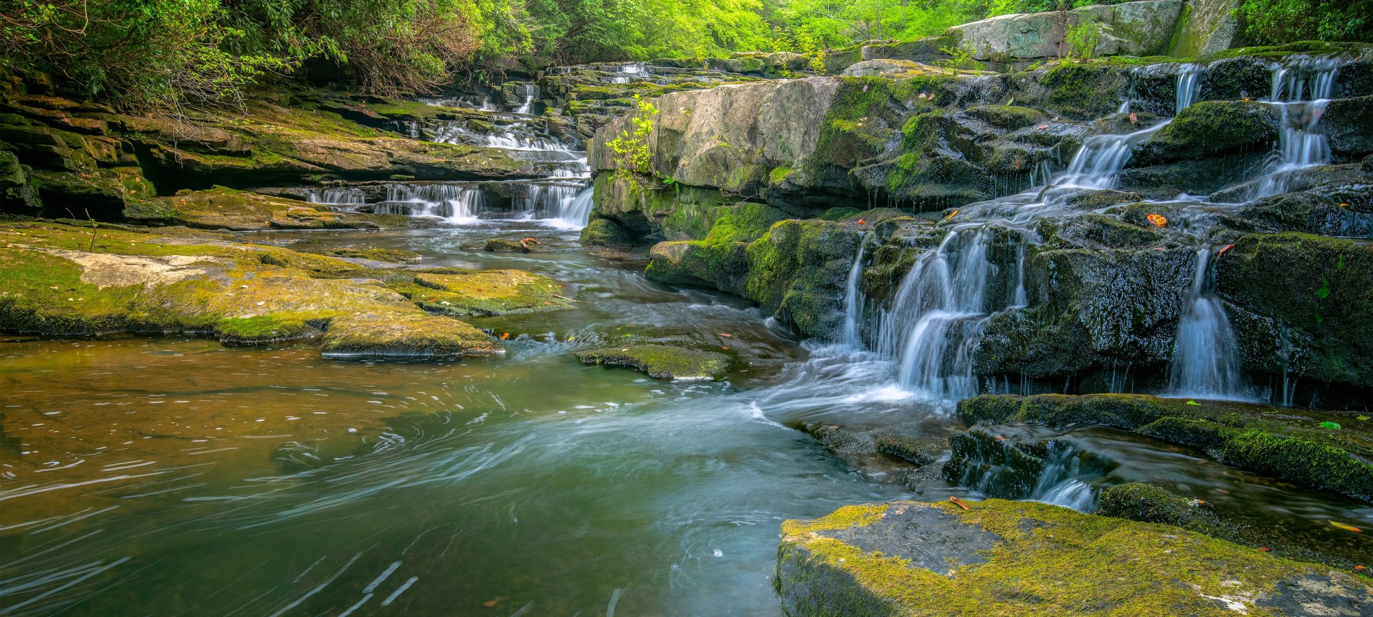 A serene waterfall cascades over moss-covered rocks into a clear, calm river, surrounded by lush green trees. The scene evokes tranquility and natural beauty.
